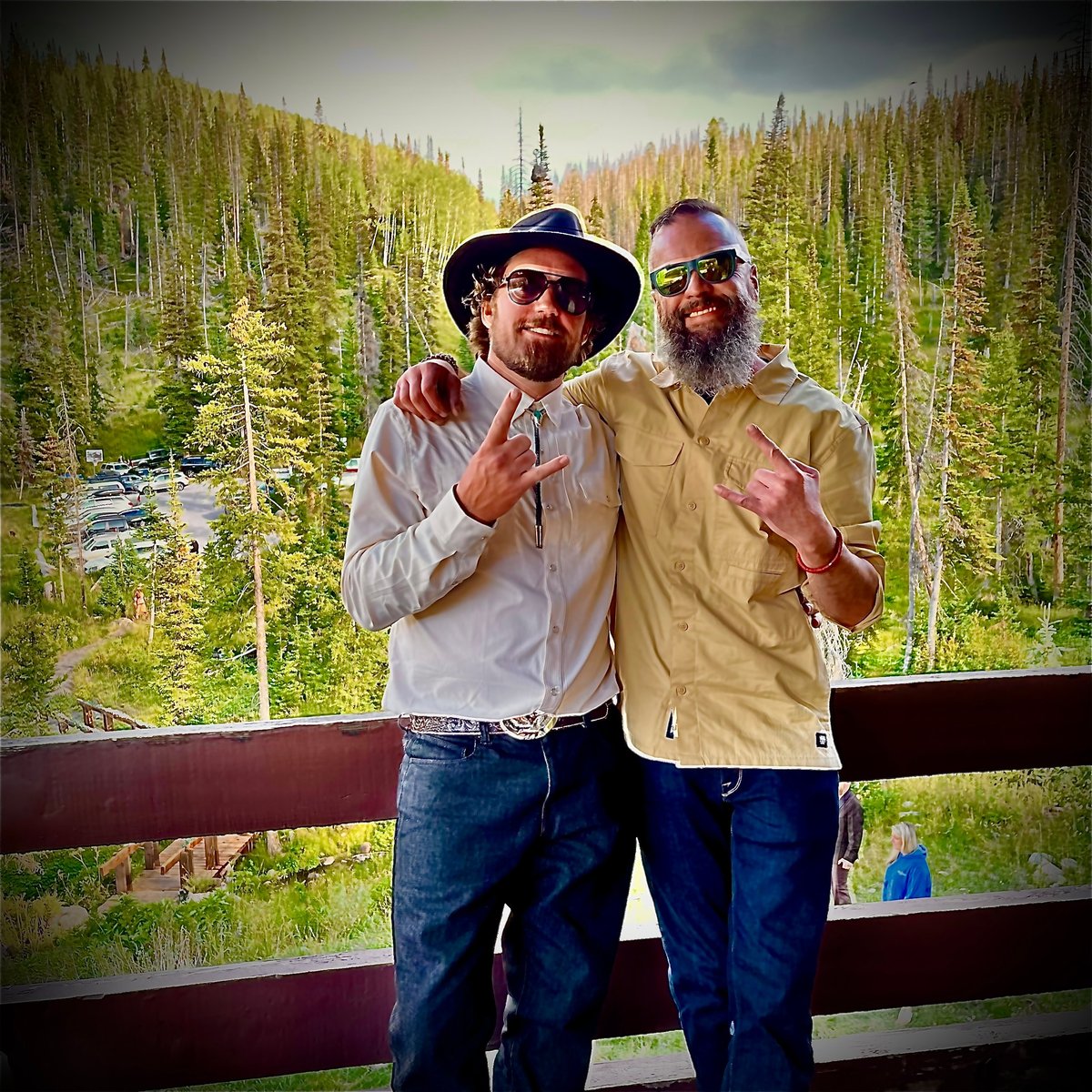 Two bearded men in hats and sunglasses pose on a railing with a vast coniferous forest landscape behind them — the TLA Promotions team