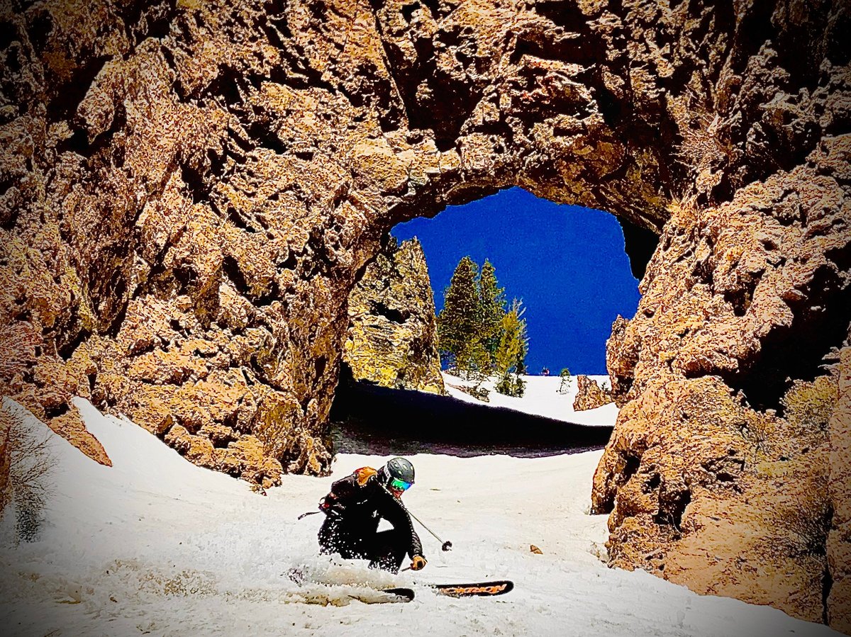 Mark skiing, sitting in snow beneath a natural rock arch opening to blue sky and distant mountain view